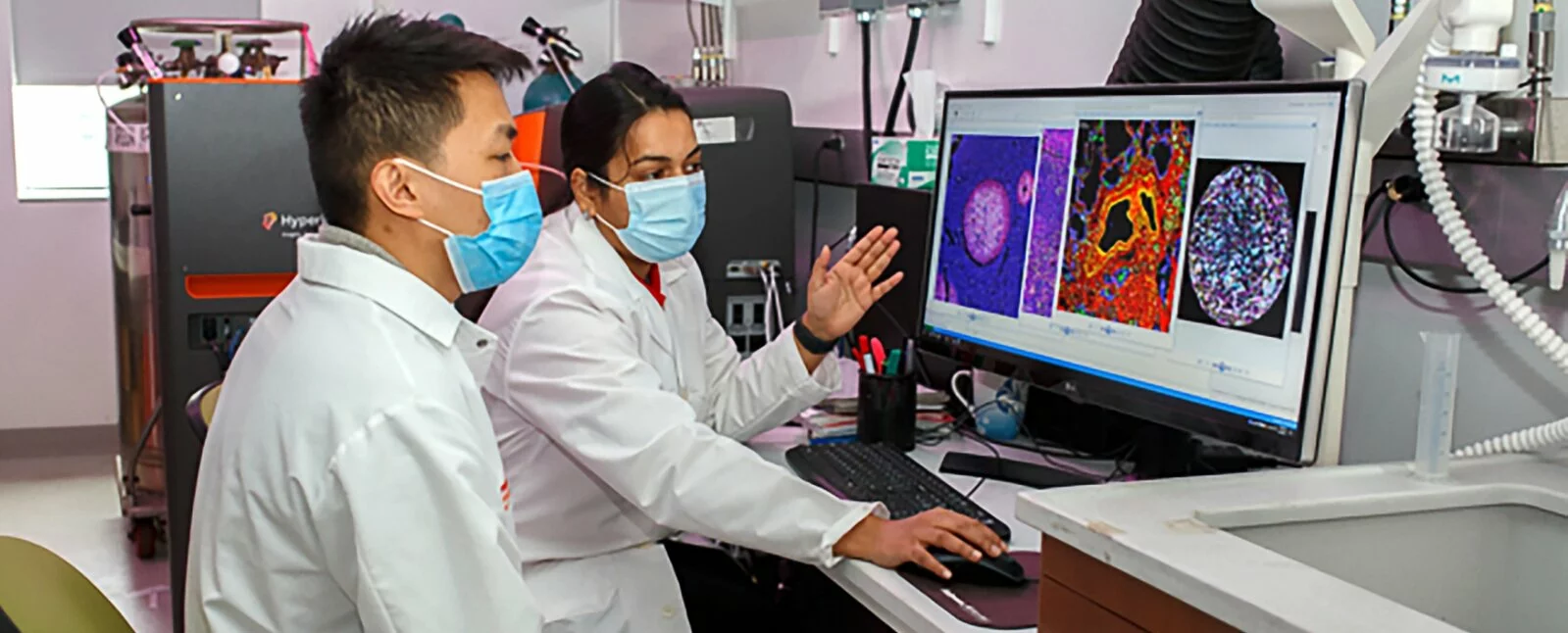 Two scientists with lab coats and masks review slides on a computer screen.