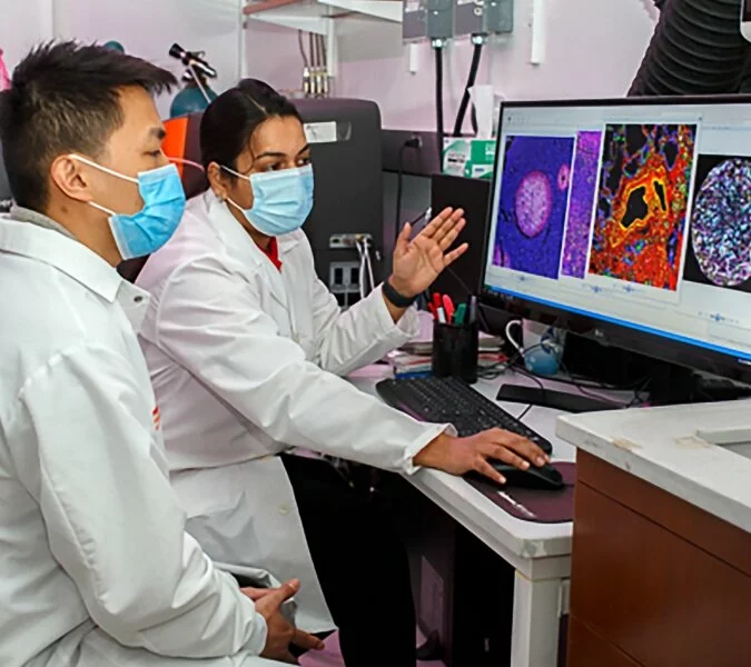 Two scientists with lab coats and masks review slides on a computer screen.