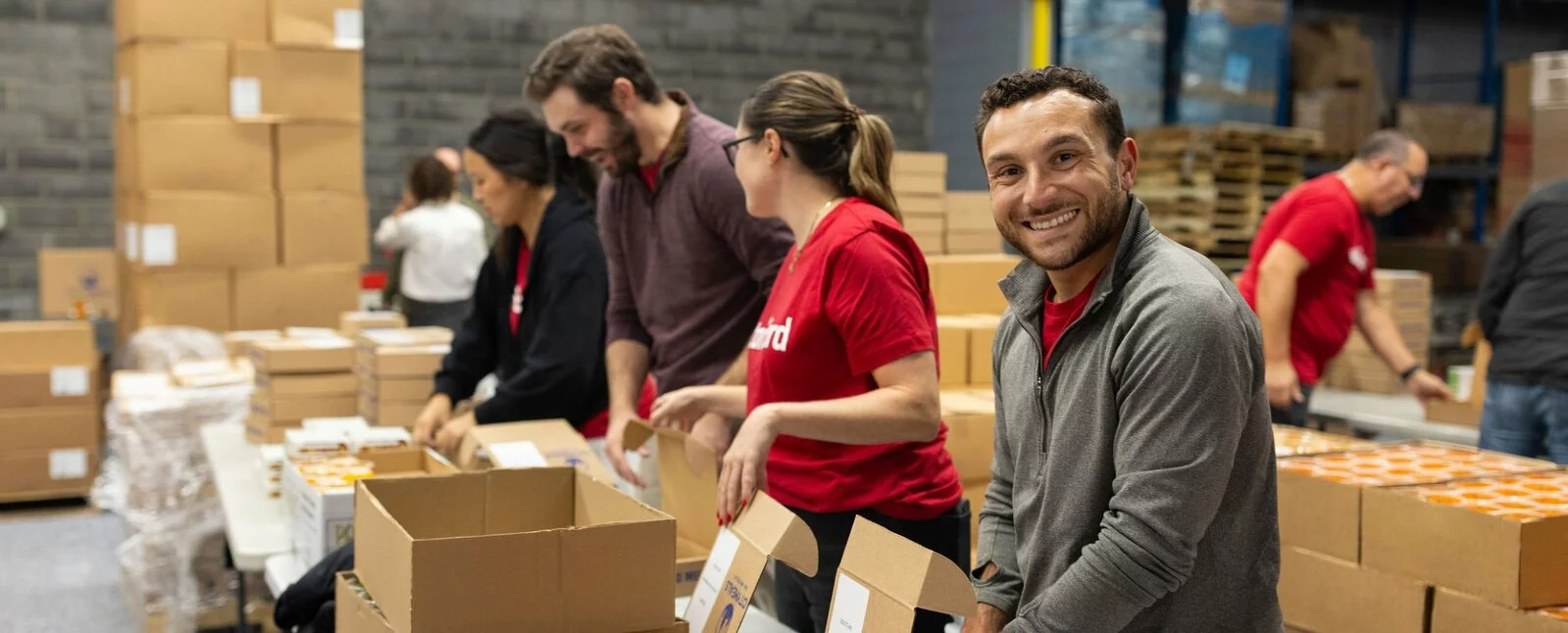 People lined up near a table with cardboard boxes put food in boxes labeled weekend meal