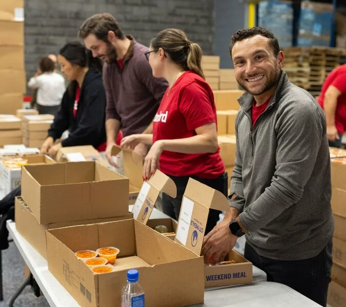 People lined up near a table with cardboard boxes put food in boxes labeled weekend meal