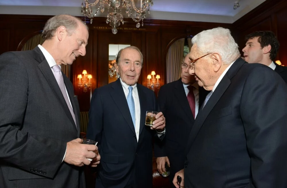 Mr. Greenberg, Richard Haass, and Dr. Henry Kissinger stand talking while other men in the background listen in.