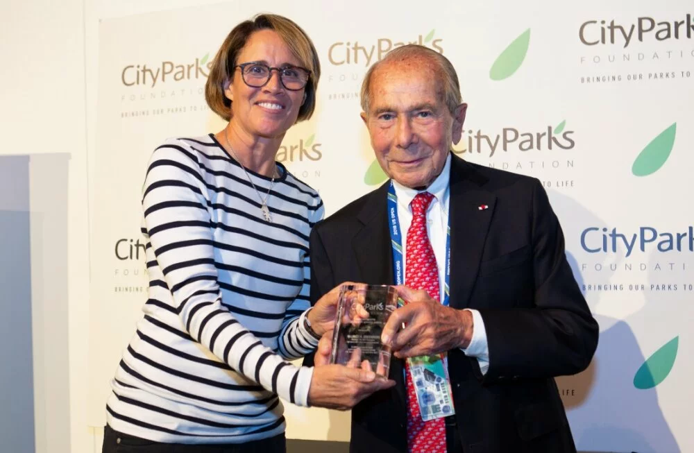 Mr. Greenberg and former tennis player, Ms. Mary Carillo stand in front of a CityParks Foundation background as Mr. Greenberg is handed a glass award from Ms. Carillo.