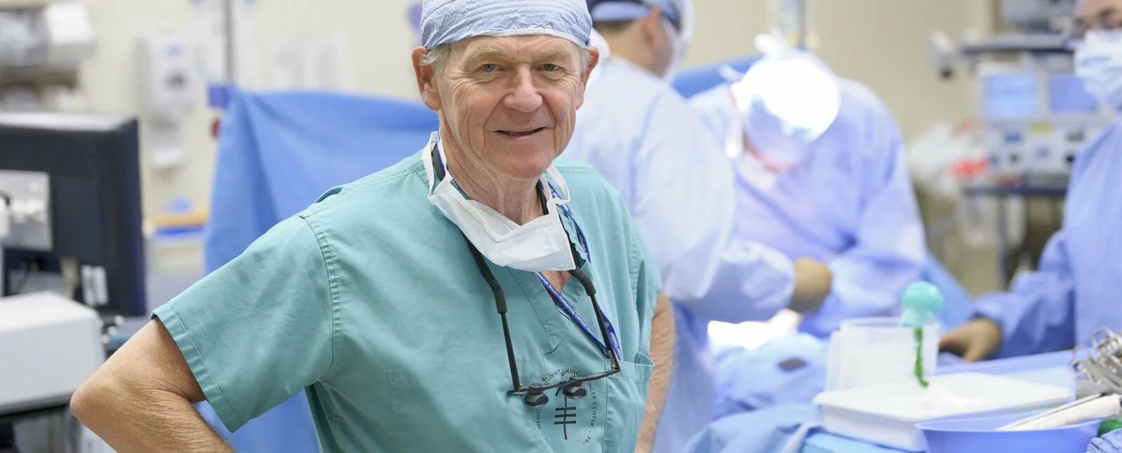 A surgeon smiles at the camera with a surgery in the background