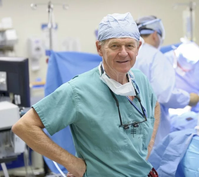 A surgeon smiles at the camera with a surgery in the background