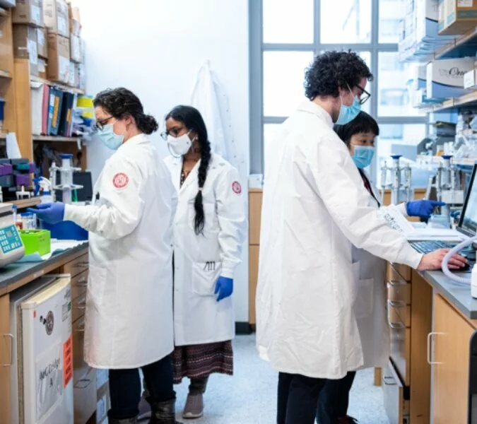 Four scientists in lab coats and masks work inside of a lab.