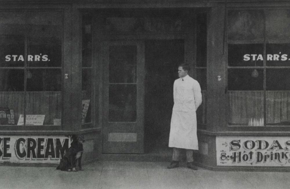 Mr. Starr, wearing a white apron, stands in front of Starr's Sodas with his dog.