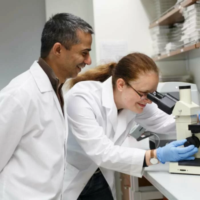 A scientist looks through microscope in a lab while another scientist smiles behind her.